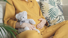 Woman Expecting Baby. Pregnant Girl Hugging Belly And Teddy Bear Toy Resting, Awaiting The Child Birth. Family Planning. Natural Pregnancy At Home