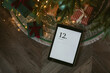 Top view of digital tablet with December calendar on screen laying with presents under Christmas tree. Holiday planning, online shopping, mockup.