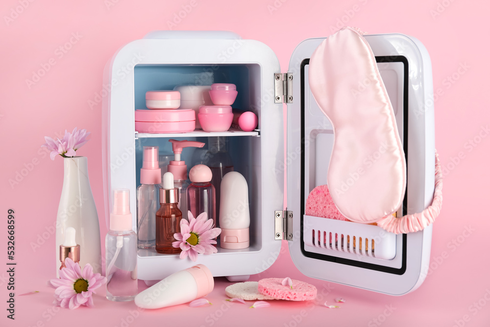 Small refrigerator with cosmetic products and flowers on pink background