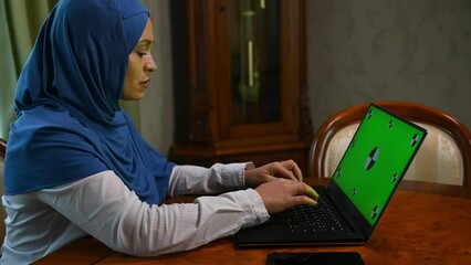 Hardworking Muslim Middle Eastern business woman in hijab, typing text on keyboard, writing computer programs while working remotely on laptop with green chroma key. Mockup screen for advertising clip