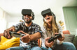 © Davide Angelini - Happy friends sitting on sofa playing video games with virtual reality glasses - Cheerful couple having fun with new trendy technology videogames - Hobby and tech concept