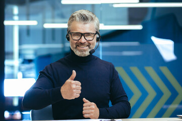 Wall Mural - Video call senior gray haired man talking to colleagues remotely using headset smiling and looking at camera giving thumbs up, mature businessman inside modern office.