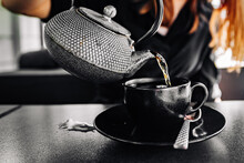 Woman Hand With Metal Teapot Pour Black Tea In Black Glass Cup In Cafe