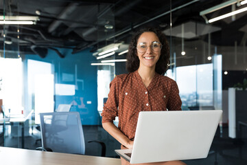Wall Mural - Portrait of young beautiful hispanic woman with laptop, business woman inside modern office building smiling and looking at camera.