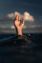 Front Of Man's Hand Reaching Out Of Water Of Blue Ocean At Sunset With Puffy White Clouds In Background, Conceptual Image With Guy's Hand Just Above Wave Symbolizing Sinking