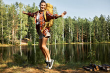 Woman Hiker With Big Yellow Backpack On Shoulders Jumping Up, Happy With Finding Best Place To Camp For Weekend, On Background With Calm Tranquil Lake And Forest On Opposite Fiver Back