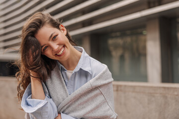 Wall Mural - Nice young caucasian girl tilting her head smiles teeth at camera outdoors. Brunette woman with wavy hair wears casual clothes. Concept of real emotions.