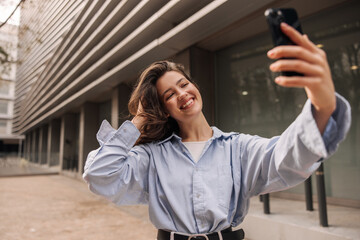 Wall Mural - Beautiful young caucasian woman with smile on face makes selfie on phone standing street. Beauty with dark wavy hair wears blue shirt. Concept of use, lifestyle