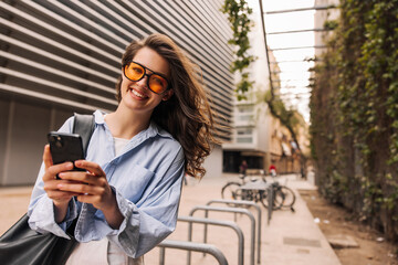 Wall Mural - Beautiful young caucasian woman in sunglasses holding smartphone outdoors. Brunette with wavy hair wears blue shirt. Concept of use