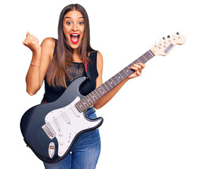 Wall Mural - Young hispanic woman playing electric guitar screaming proud, celebrating victory and success very excited with raised arms