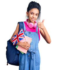 Wall Mural - Cute african american girl wearing student backpack holding united kingdom flag smiling happy and positive, thumb up doing excellent and approval sign