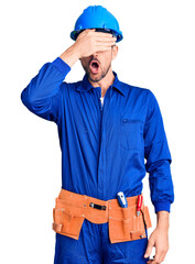 Canvas Print - Young handsome man wearing worker uniform and hardhat smiling and laughing with hand on face covering eyes for surprise. blind concept.