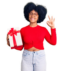 Sticker - Young african american girl holding gift smiling positive doing ok sign with hand and fingers. successful expression.