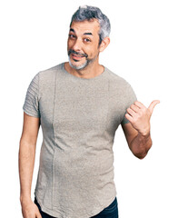 Sticker - Middle age hispanic with grey hair wearing casual grey t shirt smiling with happy face looking and pointing to the side with thumb up.