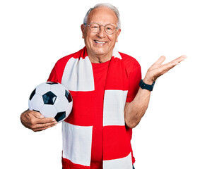 Wall Mural - Senior man with grey hair football hooligan holding ball smiling cheerful presenting and pointing with palm of hand looking at the camera.