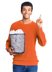 Poster - Young african amercian man holding paper bin full of crumpled papers surprised with an idea or question pointing finger with happy face, number one