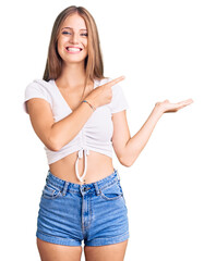 Wall Mural - Young beautiful blonde woman wearing casual white tshirt amazed and smiling to the camera while presenting with hand and pointing with finger.