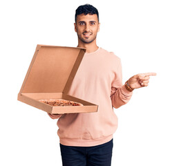 Wall Mural - Young hispanic man holding delivery pizza box smiling happy pointing with hand and finger to the side