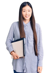 Wall Mural - Young beautiful chinese woman holding notebook looking positive and happy standing and smiling with a confident smile showing teeth