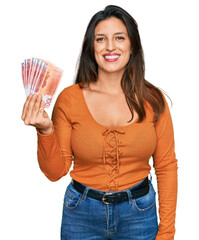 Beautiful hispanic woman holding norwegian krone banknotes looking positive and happy standing and smiling with a confident smile showing teeth