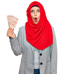 Poster - Beautiful hispanic woman wearing islamic hijab holding turkish lira banknotes scared and amazed with open mouth for surprise, disbelief face