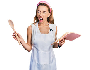 Wall Mural - Young caucasian woman wearing professional baker apron reading cooking recipe book angry and mad screaming frustrated and furious, shouting with anger. rage and aggressive concept.