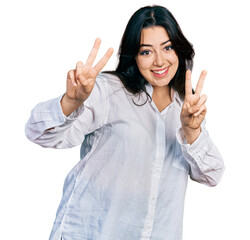 Wall Mural - Beautiful hispanic woman wearing casual white shirt smiling looking to the camera showing fingers doing victory sign. number two.