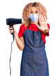 Young blonde woman with curly hair holding dryer blow wearing safety mask for coranvirus with open hand doing stop sign with serious and confident expression, defense gesture