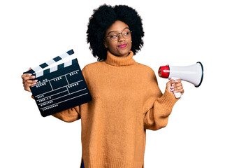 Poster - Young african american woman holding video film clapboard and megaphone smiling looking to the side and staring away thinking.