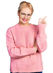 Wall Mural - Young blonde woman wearing casual clothes and glasses with a big smile on face, pointing with hand and finger to the side looking at the camera.