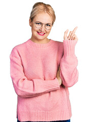 Canvas Print - Young blonde woman wearing casual clothes and glasses with a big smile on face, pointing with hand and finger to the side looking at the camera.