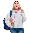 © Krakenimages.com - Hispanic woman with pink hair wearing student backpack and headphones amazed and surprised looking up and pointing with fingers and raised arms.