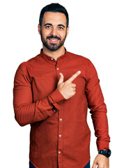 Wall Mural - Young hispanic man with beard wearing casual shirt cheerful with a smile on face pointing with hand and finger up to the side with happy and natural expression