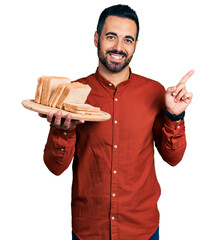 Sticker - Young hispanic man with beard holding bread smiling happy pointing with hand and finger to the side