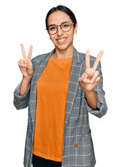 Wall Mural - Young hispanic girl wearing business jacket and glasses smiling looking to the camera showing fingers doing victory sign. number two.