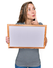 Canvas Print - Young blonde woman holding empty white board serious face thinking about question with hand on chin, thoughtful about confusing idea