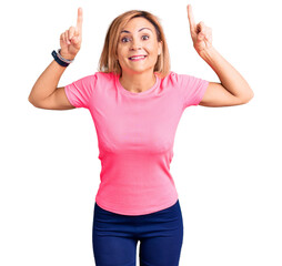 Sticker - Young blonde woman wearing sportswear smiling amazed and surprised and pointing up with fingers and raised arms.