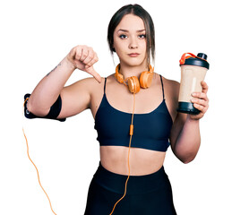 Young hispanic girl wearing sport clothes drinking a protein shake with angry face, negative sign showing dislike with thumbs down, rejection concept