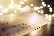 Old Sheet With Christmas Music Notes As Background Against Blurred Lights. Christmas Music Concept
