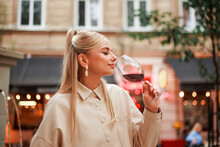 Charming Young Woman In Beige Coat On Evening Street Of An Old European City Tastes And Enjoys Red Wine