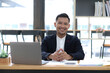 © wichayada - Portrait of a young Asian businessman smiling while using a laptop and writing down notes while sitting at his desk in a modern office