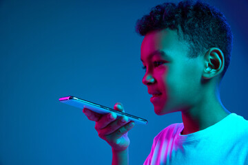 Cute little boy, kid using smartphone isolated over blue background in neon light. Education, childhood, modern lifestyle