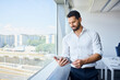 © baranq - Successful businessman using tablet at office stands by the window. Male entrepreneur in modern office looking at tablet