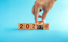 The Calendar Year 2022 Changed To 2023 With The Goal And Successful Concept. Hand Turning Wooden Cube Blocks For The Transition From 2022 To 2023 With Target Icon Sign Isolated On Blue Background.