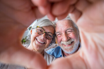 Sticker - Couple of two old and happy seniors having fun at home on the sofa doing a heart shape with their hands and fingers looking at the camera. .