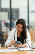 © crizzystudio - An Asian businesswoman happily uses her smartphone to respond to customer chats at a desk in a modern office.