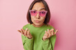 Indoor shot of Asian woman with short dark hair spreads palms feels unaware wears sunglasses and green pullover cannot decide what to do isolated over pink background. People and decision concept