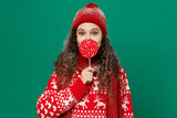 Merry young woman wearing red warm cozy knitted sweater hat posing hold cover mouth with pink lollipop candy isolated on plain dark green background. Happy New Year 2023 celebration holiday concept.