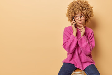 Wall Mural - Indoor shot of beautiful young woman with curly hair holds hand on chin has telephone talk looks aside with serious expression wears pink jumper and jeans poses on chair against beige background