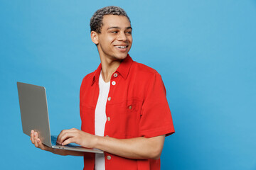 Wall Mural - Young happy fun IT man of African American ethnicity 20s he wear red shirt hold use work on laptop pc computer look aside on workspace area mock up isolated on plain pastel light blue cyan background.